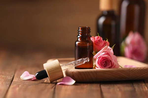 Bottle of essential rose oil and flowers on wooden table, space for text