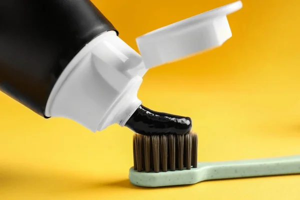 Applying charcoal toothpaste on brush against yellow background, closeup