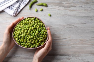 Woman holding bowl of edamame beans at light wooden table, closeup. Space for text