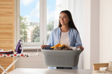 Young woman with basket full of clean laundry at table indoors