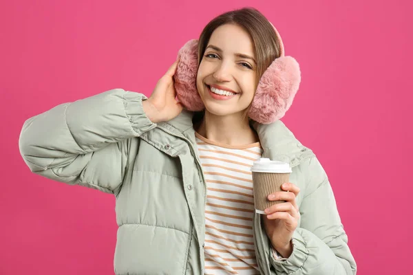 幸せな女性とともにカップのドリンク身に着けています暖かい耳飾り上のピンクの背景 — ストック写真