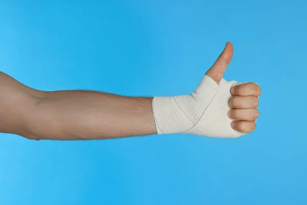 Man with hand wrapped in medical bandage showing thumbs up on light blue background, closeup