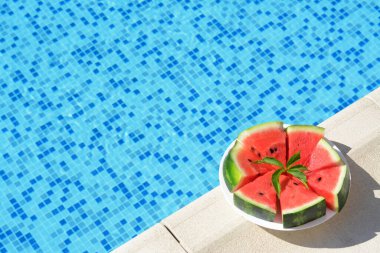 Slices of watermelon on white plate near swimming pool outdoors. Space for text