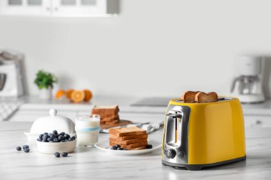 Kızarmış ekmek dilimleri, yaban mersini ve beyaz mermer masada bir bardak sütle birlikte sarı tost makinesi.