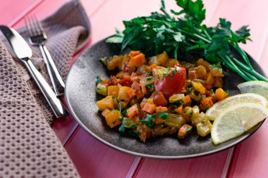 Siyah bir tabakta Ratatouille, farklı sebzeleri bir güveçte boyadı, taze maydanoz yaprakları ve limon dilimleri ile, pembe ahşap arka planda, birçok ülke için uygun fiyatlı bir yemek.