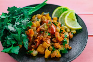 Siyah bir tabakta Ratatouille, farklı sebzeleri bir güveçte boyadı, taze maydanoz yaprakları ve limon dilimleri ile, pembe ahşap arka planda, birçok ülke için uygun fiyatlı bir yemek.