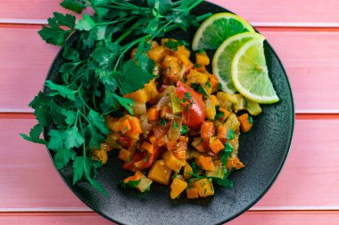 Siyah bir tabakta Ratatouille, farklı sebzeleri bir güveçte boyadı, taze maydanoz yaprakları ve limon dilimleri ile, pembe ahşap arka planda, birçok ülke için uygun fiyatlı bir yemek.