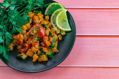 Siyah bir tabakta Ratatouille, farklı sebzeleri bir güveçte boyadı, taze maydanoz yaprakları ve limon dilimleri ile, pembe ahşap arka planda, birçok ülke için uygun fiyatlı bir yemek.