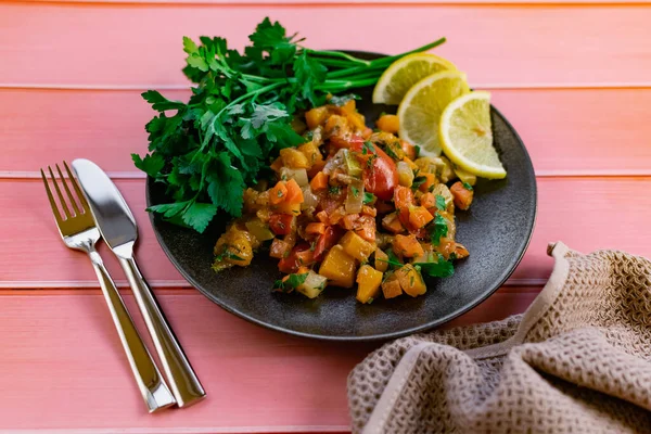 Ratatouille on a black plate, colored different vegetables in a stew, with fresh parsley leaves and lemon slices, on a pink wooden background, an affordable dish for many nations