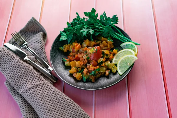 Siyah bir tabakta Ratatouille, farklı sebzeleri bir güveçte boyadı, taze maydanoz yaprakları ve limon dilimleri ile, pembe ahşap arka planda, birçok ülke için uygun fiyatlı bir yemek.