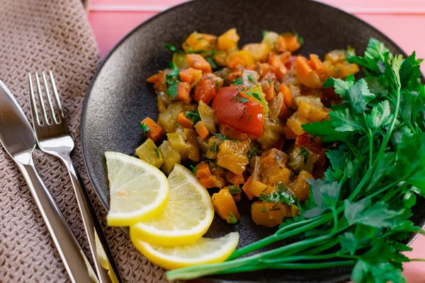 Ratatouille on a black plate, colored different vegetables in a stew, with fresh parsley leaves and lemon slices, on a pink wooden background, an affordable dish for many nations