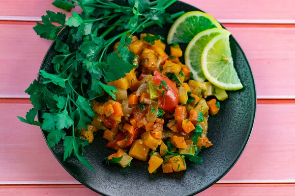 Siyah bir tabakta Ratatouille, farklı sebzeleri bir güveçte boyadı, taze maydanoz yaprakları ve limon dilimleri ile, pembe ahşap arka planda, birçok ülke için uygun fiyatlı bir yemek.