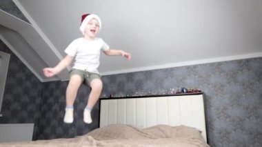 Kafasında Noel Baba şapkası olan küçük mutlu bir çocuk büyük bir yatağa atlıyor.