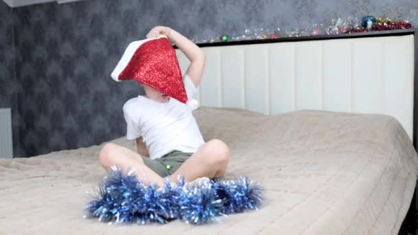 Mały chłopiec walczy z czapką Świętego Mikołaja i świecidełkiem siedzącym na dużym łóżku — Wideo stockowe