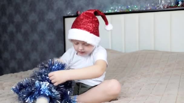 Een kleine jongen knoeit met een kerstman pet en knutselen zittend op een groot bed — Stockvideo