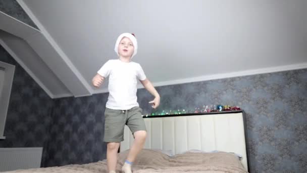 En glad liten pojke med en tomte mössa på huvudet hoppar på en stor säng — Stockvideo
