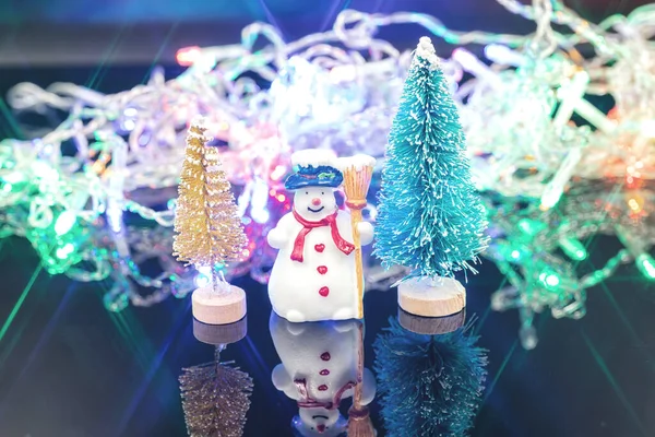 Un muñeco de nieve se para en un árbol de Navidad contra una guirnalda destellante. — Foto de Stock