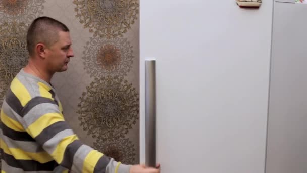 Мужчина открывает холодильник на кухне и исчезает в нем, дверь закрывается — стоковое видео