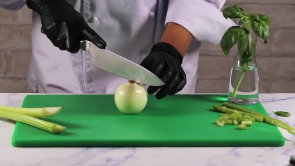 Donna in grembiule bianco in guanti neri taglia cipolla su un tagliere verde con un grande coltello — Video Stock