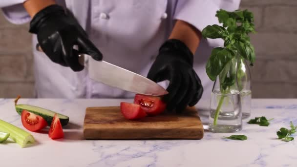 Mãos femininas em avental branco luvas pretas cortando tomate na placa verde, atrás de legumes frescos. — Vídeo de Stock