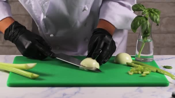 Frau in weißer Schürze und schwarzen Handschuhen schneidet mit einem großen Messer Zwiebeln auf einem grünen Schneidebrett — Stockvideo