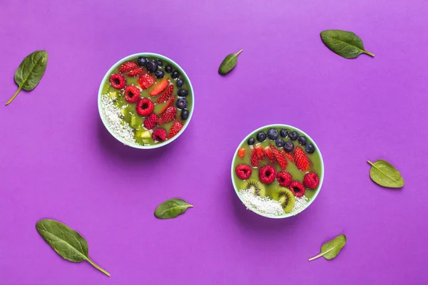 Tazón de batido con bayas frescas y frutas en plato azul sobre violeta con hojas de espinaca Fotos De Stock