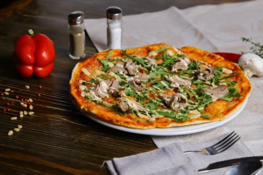 Ahşap bir masada, mantarlı pizza ahşap bir masada, pizza düz bir tabakta keten peçetenin üzerinde. Bir takım cihazlar. Otlu mantarlar telaffuz edilir..