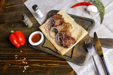 Koyu renk ahşap bir masanın üstünde, keten bir peçetenin üzerinde, kesim tahtasının üzerinde, üzerinde kırmızı soğan halkalarından yapılmış bir şiş kebabı, bir tencerede sos, kırmızı biber sürüsü, çatal bıçak takımı olan bir Ermeni lahanası var..