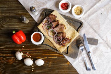 Koyu renk ahşap bir masanın üstünde, keten bir peçetenin üzerinde, kesim tahtasının üzerinde, üzerinde kırmızı soğan halkalarından yapılmış bir şiş kebabı, bir tencerede sos, kırmızı biber sürüsü, çatal bıçak takımı olan bir Ermeni lahanası var..