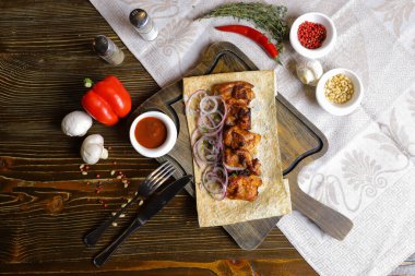 Koyu bir masa üstünde, açık keten bir peçete, pide ekmeğindeki kesme tahtasının üzerinde kırmızı soğan halkaları, çatal bıçak takımı, bir çift tuz ve biber, bir tutam acı biber, baharatlar, çeşitli renklerde serpilmiş hazır bir şiş kebap var. 