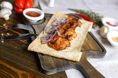 Koyu bir masa üstünde, açık keten bir peçete, pide ekmeğindeki kesme tahtasının üzerinde kırmızı soğan halkaları, çatal bıçak takımı, bir çift tuz ve biber, bir tutam acı biber, baharatlar, çeşitli renklerde serpilmiş hazır bir şiş kebap var. 