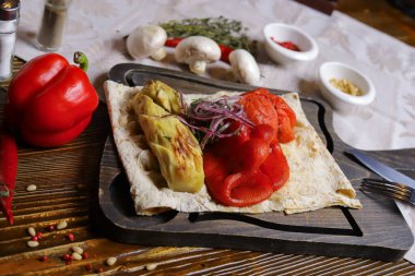 Pita ekmeği ızgara sebze kabağı, kırmızı biber, koyu renk kesim tahtasında derisiz domates, siyah masa üstünde keten peçete ile servis edilir. Bir çift biber ve tuz, çatal bıçak takımı, beyaz mantar, bütün kırmızı biber, acı biber, ro