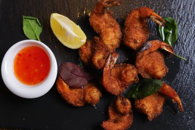 Karides Tempura kralı karidesleri, dilimlenmiş limon, yeşil fesleğen yaprakları siyah bir taş standında servis edilir. Çatal bıçak takımı, acı biber kabukları, kasedeki baharatlar, beyaz mantarlar..