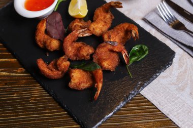 Karides Tempura kralı karidesleri, dilimlenmiş limon, yeşil fesleğen yaprakları siyah bir taş standında servis edilir. Çatal bıçak takımı, acı biber kabukları, kasedeki baharatlar, beyaz mantarlar..