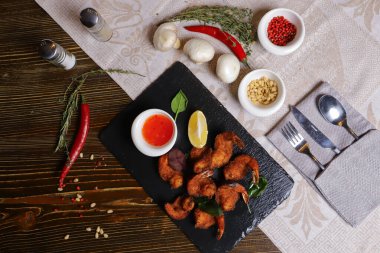 Karides Tempura kralı karidesleri, dilimlenmiş limon, yeşil fesleğen yaprakları siyah bir taş standında servis edilir. Çatal bıçak takımı, acı biber kabukları, kasedeki baharatlar, beyaz mantarlar..