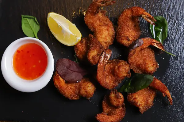 Karides Tempura kralı karidesleri, dilimlenmiş limon, yeşil fesleğen yaprakları siyah bir taş standında servis edilir. Çatal bıçak takımı, acı biber kabukları, kasedeki baharatlar, beyaz mantarlar..