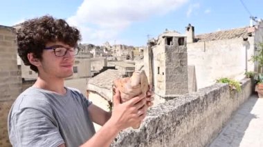Matera, Basilicata, İtalya. Ağustos 2021. Kıvırcık saçlı ve gözlüklü sevimli beyaz bir çocuğun görüntüsü. Tarihi merkezde, yerel bir spesiyal olan Matera ekmeğinin büyük bir somununu ısırıyormuş gibi yapıyor..