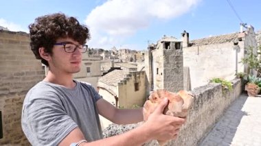 Matera, Basilicata, İtalya. Ağustos 2021. Kıvırcık saçlı ve gözlüklü beyaz bir çocuğun yavaş çekim görüntüleri. Tarihi merkezde büyük bir somun Matera ekmeğini ısırma simülasyonu yapıyor..