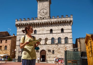 Montepulciano, Toskana, İtalya. Ağustos 2020. Belediyenin meydanında elinde turist haritası olan beyaz bir kadın etrafa bakıyor ve kadına odaklanıyor. Arkasında belediye binası var..