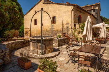Bagno Vignoni, Toskana, İtalya. Ağustos 2020. Köyün merkezinde açık masaları ve şemsiyeleri olan bir restoran var. Eski çağlardan kalma bir kuyu içeriğe birçok cazibe katıyor..