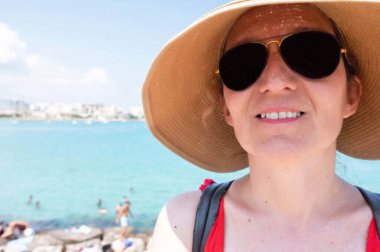 Güneş gözlüklü ve yaz şapkalı orta yaşlı bir kadın. Etrafına bakıyor ve Otranto şehrinin tadını çıkarıyor. Arka plan bulanık. Güzel bir yaz günü.