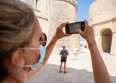 Otranto, Puglia, İtalya. Ağustos 2021. Güneş gözlüklü ve koronavirüs maskeli orta yaşlı bir kadın kalenin duvarlarında akıllı telefonuyla onun fotoğrafını çekiyor..