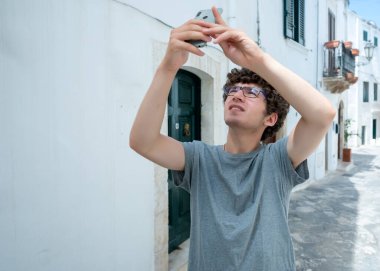 Ostuni, Puglia, İtalya. Ağustos 2021. Genç bir Kafkasyalı, köyün tarihi merkezinin beyaz sokaklarından birinde cep telefonuyla fotoğraf çekmekle meşgul. Bulanık arkaplan.