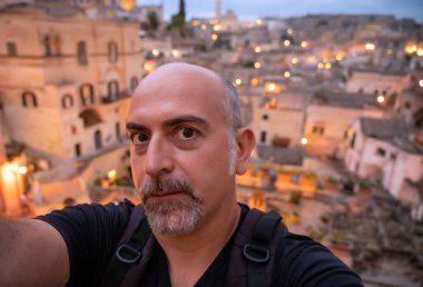 Matera, Basilicata, İtalya. Orta yaşlı bir adam arkasında tarihi bir merkez olan Matera taşlarıyla selfie çeker. Mavi saat, şehrin ışıkları açık..
