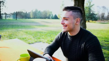 Gündelik giysiler içinde genç ve çekici bir ofis adamının portresi. Konuşuyor ve çalışmaya odaklanıyor. Parktaki bir masadan tabletle uzaktan kumandayla çalışıyor.