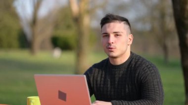 Gayri resmi giyinen, konuşan, gülümseyen ve işine odaklanan çekici genç ofis çalışanının portresi. Parktaki bir masadan dizüstü bilgisayarla uzaktan kumandalı olarak çalışıyor.