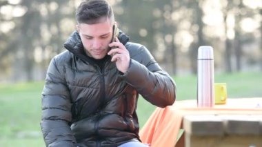 Gayri resmi giyinmiş genç ofis çalışanının portresi, cep telefonuyla konuşuyor, gülümsüyor ve dizüstü bilgisayarında daktilo yazıyor. Masanın üzerinde bir fincan kahve ve kahve. Parkta uzaktan çalışıyorum..