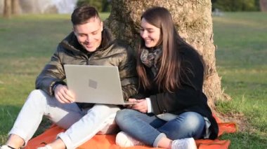 Gündelik giyinen güzel bir çift dışarıda dizüstü bilgisayarla eğleniyor. Bir ağacın dibinde otururken güneşli bir bahar gününde doğanın tadını çıkarırsın. Uzak takım çalışması kavramı.