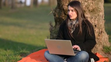 Dizüstü bilgisayarda çalışan güzel, genç esmer kadın parktaki bir ağacın dibinde oturuyor. Ara sıra etrafına bakar ve klavyede yazarken konuşur. Uzaktan çalışma konsepti. Boşluğu kopyala.