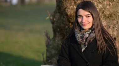 Dizüstü bilgisayarda çalışan güzel bir esmer kadının portresi parktaki bir ağacın dibinde oturuyor. Kameraya gülümsüyor ve sonra klavyeye yazarken. Uzak çalışma konsepti.